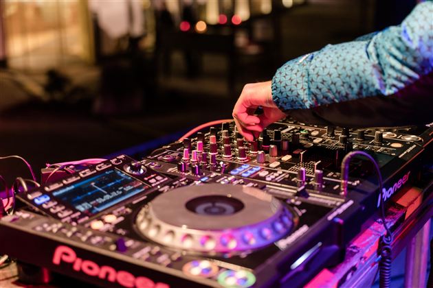 Ein DJ-Pult mit einem DJ, der an den Reglern und Knöpfen arbeitet. Bunte Lichter und eine festliche Atmosphäre sind im Hintergrund sichtbar.