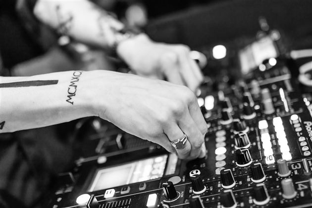 Ein DJ steht am Mischpult und steuert die Musik. Die Hände sind aktiv und fokussiert auf die Regler.