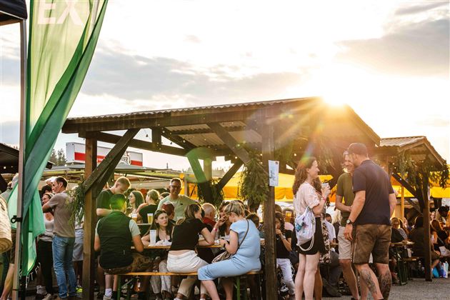Ein belebter Markt mit vielen Menschen, die an Tischen sitzen und essen. Die Sonne scheint und sorgt für eine angenehme Atmosphäre.