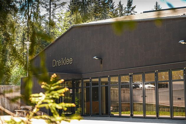 En moderne bygning med inskriptionen "Dreiklee" og store vinduer. Omgivet af træer og grønt område.