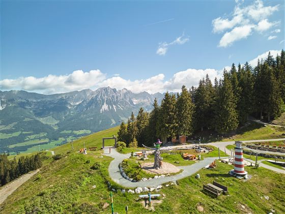 Eine wunderschöne Berglandschaft mit Wiesen und Bäumen. Im Vordergrund sind verschiedene Freizeitangebote und Skulpturen zu sehen.