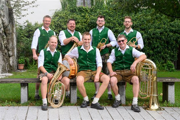 Eine Musikergruppe sitzt im Freien auf einer Bank. Sie tragen grüne Westen und spielen verschiedene Blasinstrumente.