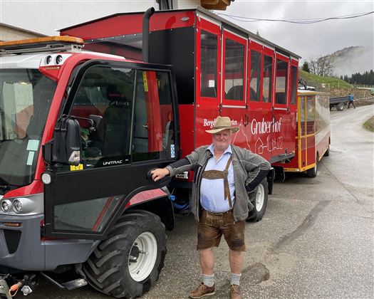 Ein Mann in traditioneller bayerischer Tracht steht neben einem Traktor mit einem roten Anhänger. Im Hintergrund sind sanfte Hügel und bewölkter Himmel zu sehen.