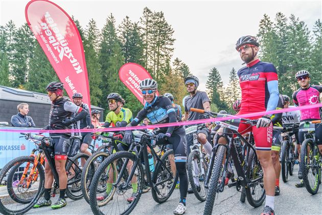 Skupina sportovních cyklistů stojí na startu mountainbikového závodu. V pozadí jsou stromy a bannery s logy.