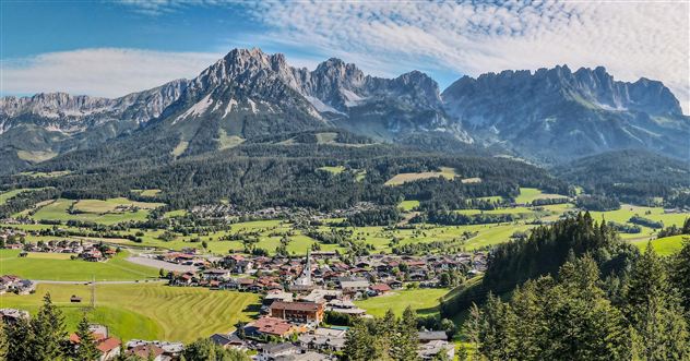 Eine beeindruckende Berglandschaft mit grünen Wiesen und einem klaren Himmel. Im Vordergrund sind kleine Häuser und geschäftige Felder zu sehen.