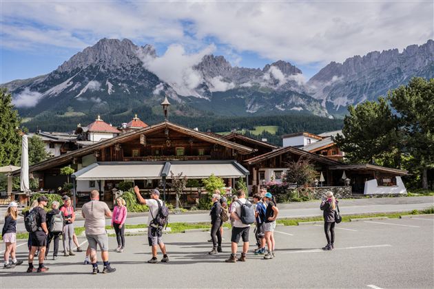 Eine Gruppe von Menschen steht vor einem traditionellen Holzhaus in den Bergen. Im Hintergrund sind majestätische Berge mit Wolken zu sehen.