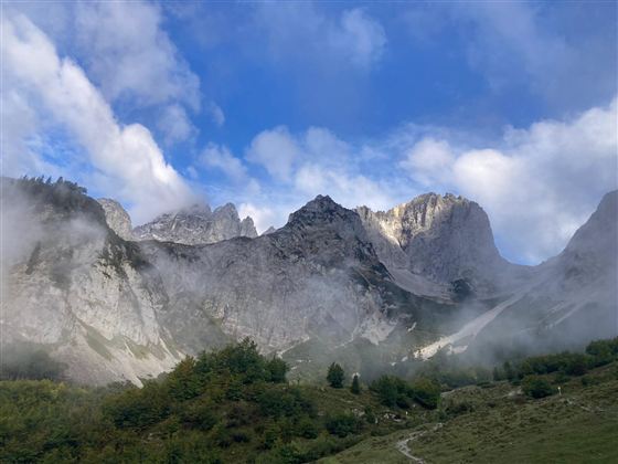Ein beeindruckendes Bergpanorama mit felsigen Gipfeln und leichten Nebelschwaden. Der Himmel ist klar mit einigen Wolken.