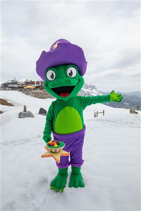 Een groene mascotte met een paarse hoed staat in de sneeuw en glimlacht. Hij houdt een schaal met eten in de hand en wijst met de andere hand naar de omgeving.