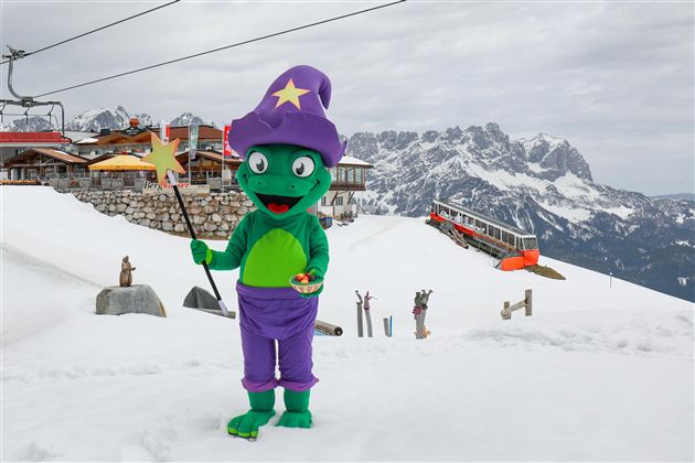Een groene tovenaar in een kleurrijk kostuum staat in de sneeuw voor een berglandschap. Op de achtergrond zijn een skilerp en een berghut te zien.
