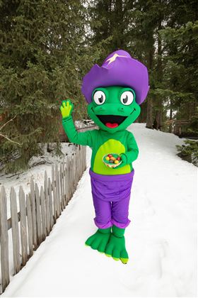 Een kleurrijk mascotte staat in de sneeuw en zwaait. Het draagt een paarse hoed en houdt een kom met kleurrijke snoepjes vast.