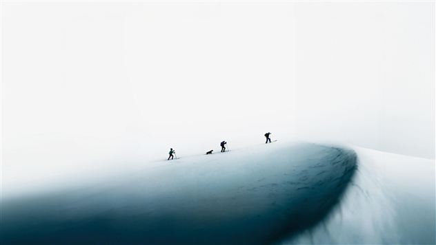 Eine Gruppe von Skifahrern wandert auf einem schneebedeckten Berg. Der Himmel ist hell, und die Landschaft wirkt minimalistisch.