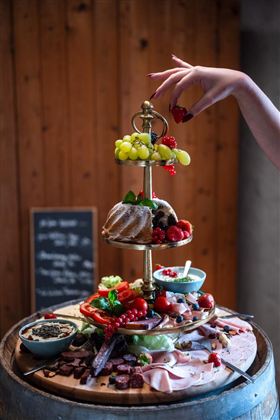 Dekorativní bufet s různými masovými a sýrovými talíři, čerstvým ovocem a barevnou zeleninou. Ruka přidává jahodu, zatímco rustikální pozadí vytváří příjemnou atmosféru.