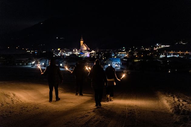 A group of people with torches walks through a snow-covered landscape at night. In the background, a city with a church is glowing.