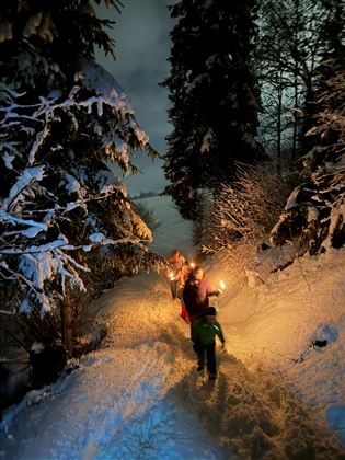 Eine Gruppe von Menschen geht mit Fackeln durch eine schneebedeckte Landschaft in der Dämmerung. Im Hintergrund sind beleuchtete Häuser sichtbar.