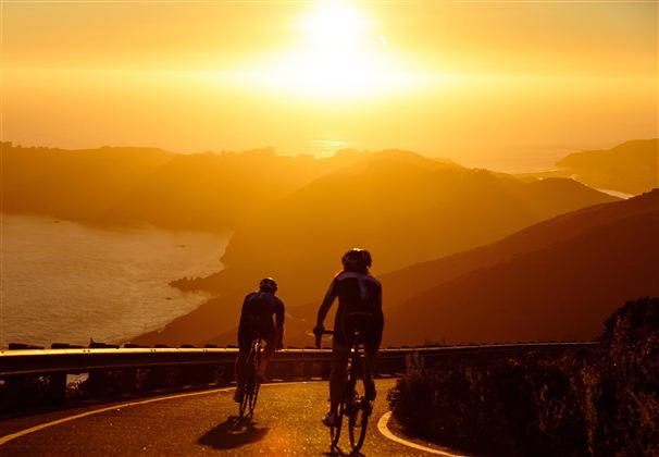 Zwei Radfahrer fahren einen kurvenreichen Weg entlang einer Küste bei Sonnenuntergang. Die Szenerie ist von warmem Licht und sanften Hügeln umgeben.
