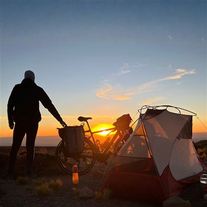 To cyklister kører ad en snoet vej langs en kyst ved solnedgang. Scenen er omgivet af varmt lys og bløde bakker.