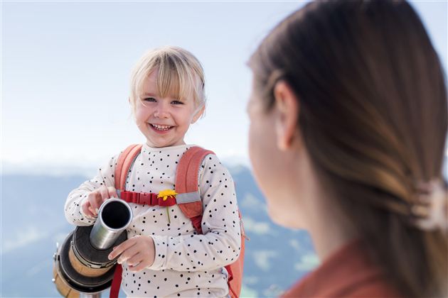 En smilende pige holder en kande og ser glad ud. I baggrunden ses bjerge og en klar himmel.