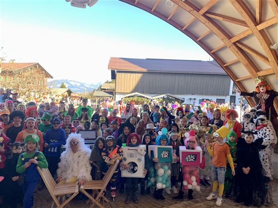 A large group of children in colorful costumes gathers under a wooden roof. In the background, mountains and a clear sky are visible.