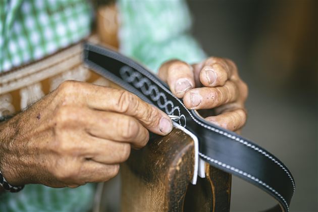 Ein Handwerker arbeitet sorgfältig an einem schwarzen Lederstück. Die Hände sind gekennzeichnet von der Handarbeit und zeigen viel Geschick.