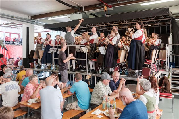 Eine Blaskapelle spielt auf einer Bühne, während das Publikum an Tischen sitzt. Die Menschen genießen das Essen und die Musik in einer geselligen Atmosphäre.