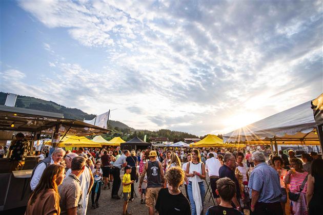 Ein lebendiger Markt mit vielen Menschen und bunten Ständen. Die Sonne scheint am Himmel und schafft eine fröhliche Atmosphäre.