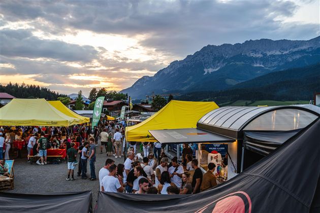Eine belebte Veranstaltung mit vielen Menschen und bunten Zelten. Im Hintergrund sind imposante Berge und ein schöner Sonnenuntergang zu sehen.
