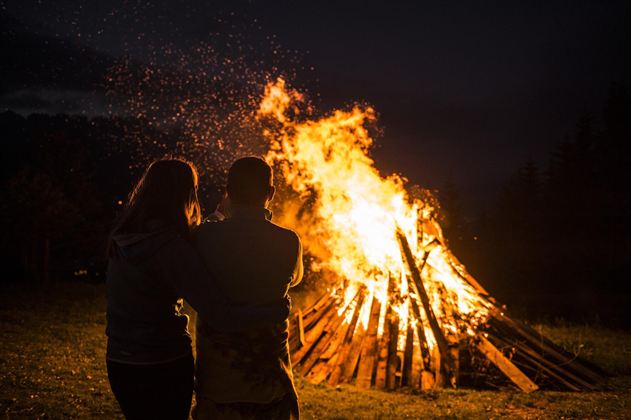 Ein Lagerfeuer in der Nacht strahlt warmes Licht aus. Zwei Personen stehen nah beieinander und schauen dem Feuer zu.