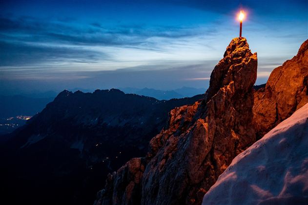 Ein beeindruckender Berghorizont bei Dämmerung, beleuchtet von einer Kerze auf einem hohen Felsen. Die Szenerie vermittelt eine ruhige und mystische Atmosphäre.