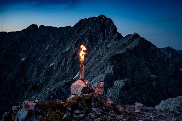Eine Fackel brennt auf einem felsigen Berggipfel in der Dämmerung. Die beeindruckende Berglandschaft bildet den Hintergrund.