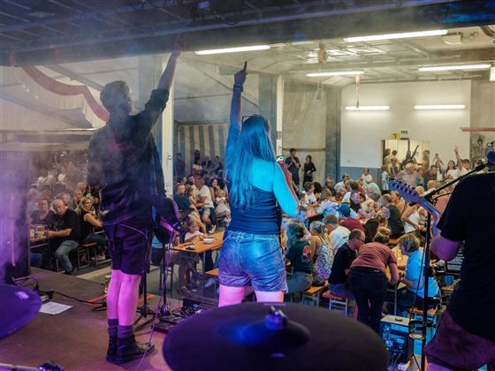 Eine Band spielt auf einer Bühne vor einem begeisterten Publikum. Die Zuschauer heben ihre Hände und feiern die Musik.