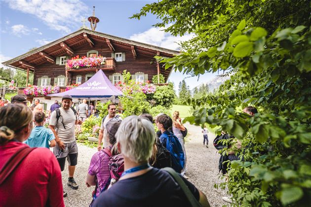 Eine Gruppe von Menschen steht vor einem schönen, traditionellen Holzhaus mit blühenden Balkonen. Der Himmel ist blau und die Umgebung ist grün und einladend.