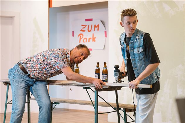 Two men are standing at a table in a room. One is leaning over the table while the other is holding a camera and looking into the distance.