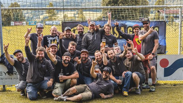 Eine Gruppe von Personen steht vor einem Tor und feiert. Sie tragen lässige Kleidung und halten Bier in den Händen.