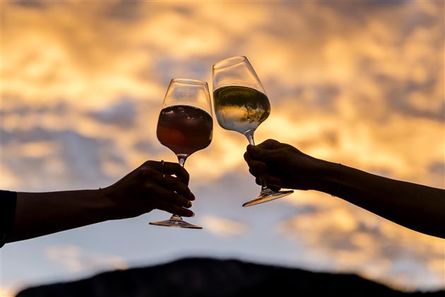 Zwei Gläser werden zum Anstoßen erhoben, silhouettiert vor einem farbenfrohen Sonnenuntergang. Die Szene vermittelt eine festliche und gesellige Stimmung.