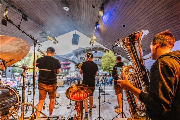 Eine Band spielt auf einer Bühne vor einem Publikum. Im Vordergrund sind Musiker mit Blasinstrumenten zu sehen.