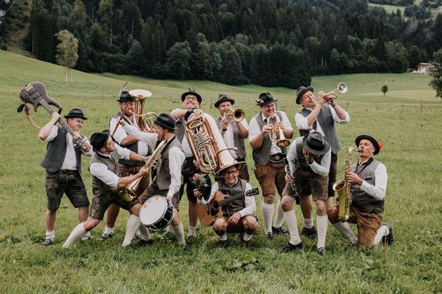 En gruppe af musikere i traditionelt tøj spiller glade melodier på en grøn eng. I baggrunden ses træer og bakker.