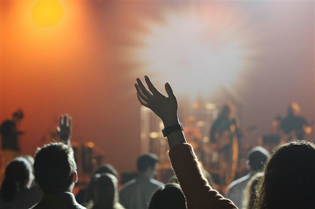 Eine Menschenmenge bei einem Konzert, die die Hände in die Luft hebt. Die Bühne ist in warmen Farben beleuchtet.