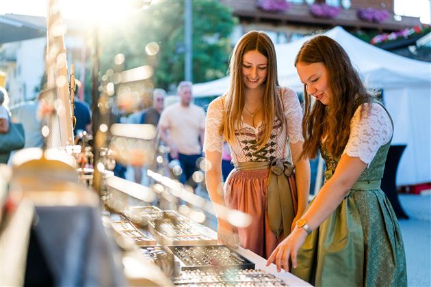 Zwei Frauen in traditionellen Kleidern schauen sich Schmuck auf einem Markt an. Die Sonne scheint, und im Hintergrund sind andere Besucher zu sehen.