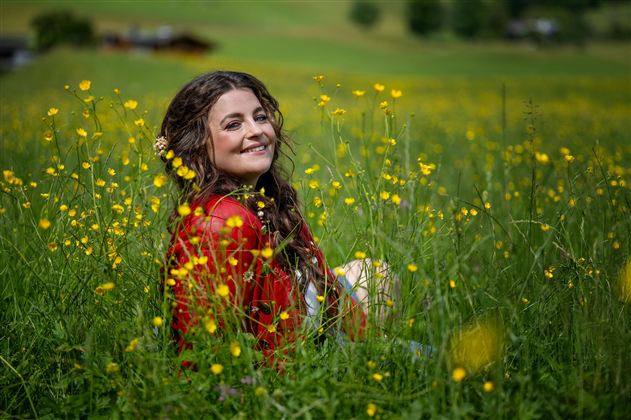 Eine lächelnde Frau sitzt in einem blühenden Feld mit gelben Blumen. Sie trägt ein rotes Kleid und genießt die Natur.
