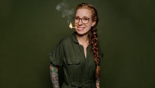 Eine lächelnde Frau mit langen, geflochtenen Haaren und Brille steht vor einem grünen Hintergrund. Sie hat Tattoos auf ihren Armen und raucht eine Zigarette.