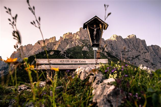 Eine malerische Berglandschaft mit einer Holztafel und einer kleinen Kapelle. Bunte Blumen wachsen im Vordergrund, während die imposing Felsen im Hintergrund sichtbar sind.