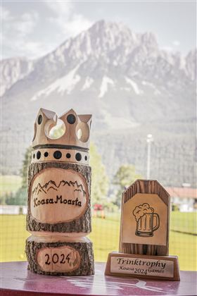 Zwei Holzpokale stehen vor einer beeindruckenden Berglandschaft. Der größere Pokal hat das Datum 2024 und die Aufschrift "Koasa Moasta".