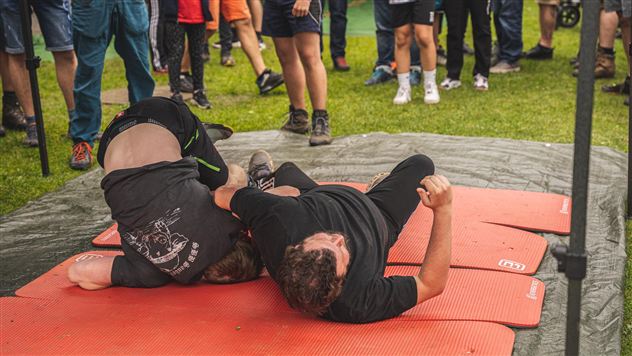 Zwei Kämpfer liegen auf roten Matten und sind im Wettkampf miteinander beschäftigt. Eine Gruppe von Zuschauern beobachtet das Geschehen im Hintergrund.