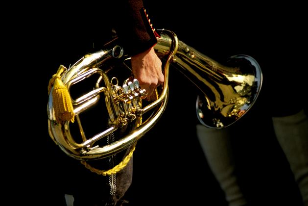 Een muzikant houdt een glanzend, gouden posthoorn in de hand. De donkere achtergrondkleur geeft het instrument extra nadruk.