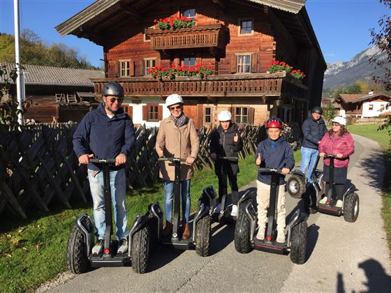 Eine Gruppe von Menschen fährt auf Segways auf einem schmalen Weg. Im Hintergrund ist ein traditionelles Holzhaus mit Blumenkästen zu sehen.