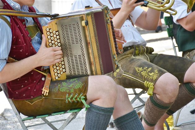 Eine Gruppe von Musikern spielt auf einem traditionellen Akkordeon. Sie tragen bayerische Trachten mit Lederhosen und sitzen im Freien.