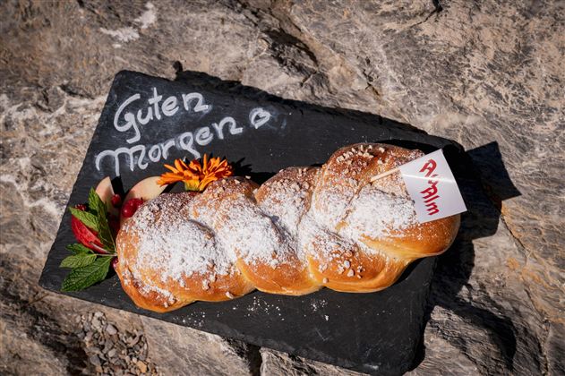 Ein frisch gebackenes, geflochtenes Brot mit Puderzucker auf einem schwarzen Steinuntergrund. Daneben steht "Guten Morgen" auf dem Stein, dekoriert mit bunten Blumen.