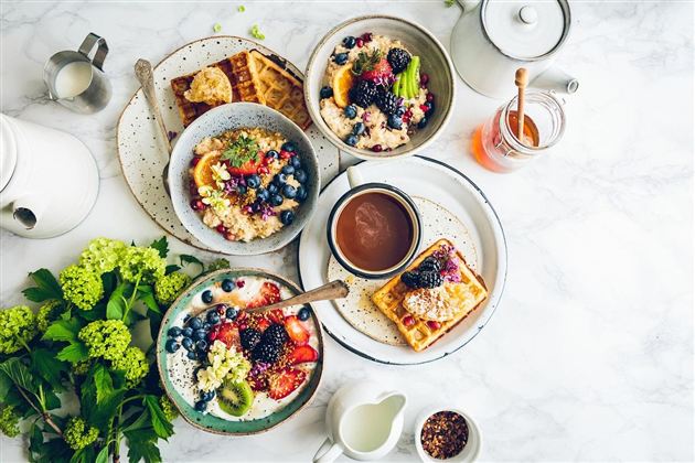 Ein schönes Frühstückstisch-Arrangement mit Waffeln, Joghurt und verschiedenen Beeren. Dazu gibt es frische Früchte und einen Krug mit Honig.