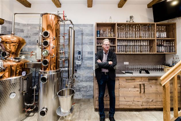A modern distillery with a copper kettle and wooden shelves. A man stands relaxed next to the equipment.