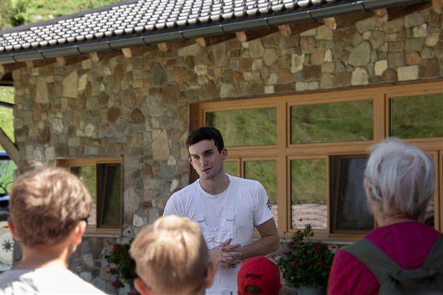 Ein junger Mann spricht zu einer Gruppe von Menschen vor einem Steinhaus. Die Zuhörer sind aufmerksam und scheinen interessiert zu sein.